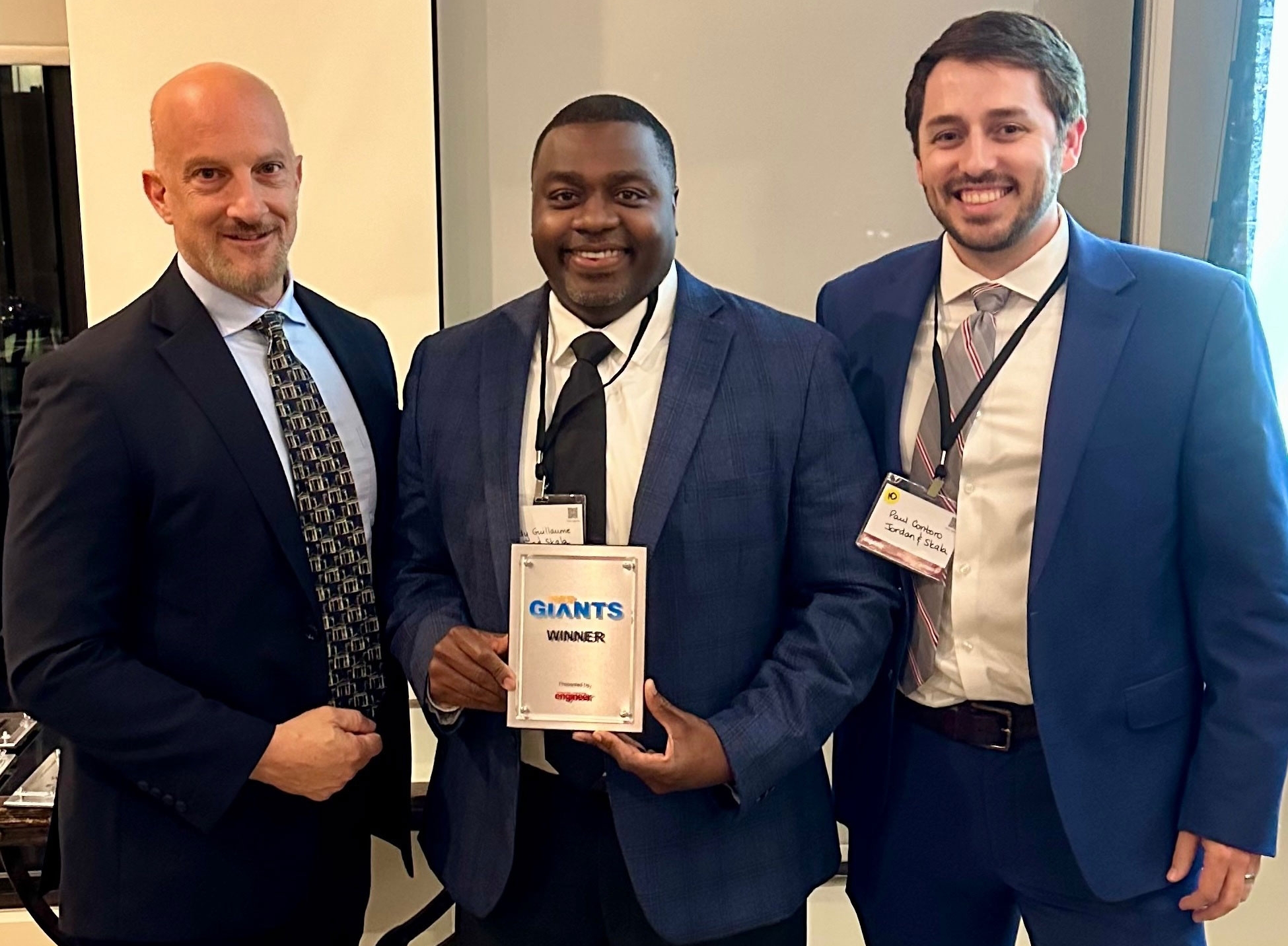 Three men standing in a line with the middle man, Kendy Guillaume, holding an award plaque.